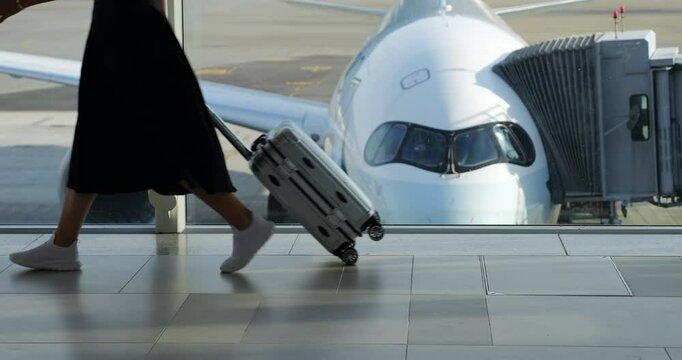 Woman with small wheeled suitcase walks through terminal past window, heading to boarding gate. Slow-motion shot shows her legs and suitcase, with modern airplane seen blurred in background.