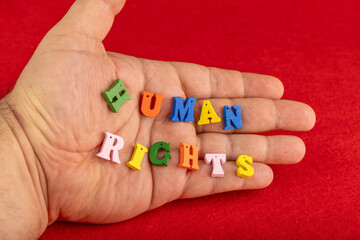 Fototapeta premium Human rights spelled out in colorful wooden letters, delicately held in a hand. The red background highlights the importance of equality and freedom