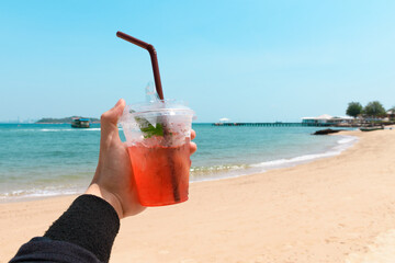 Hand holding strawberry lemon soda in cocktail glass over turquoise sea and beautiful beach against blue sky background. Hand holding a glass of fruit drink. Vacation and Holiday concept.