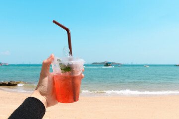 Hand holding strawberry lemon soda in cocktail glass over turquoise sea and beautiful beach against blue sky background. Hand holding a glass of fruit drink. Vacation and Holiday concept.