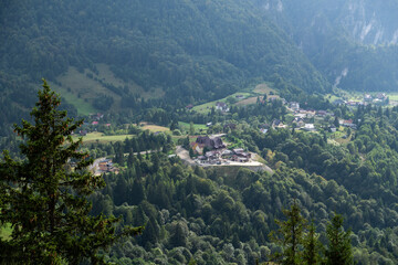 A picturesque village nestled in a lush, green valley surrounded by dense forests and rolling hills. The view from above captures scattered houses, open fields, and a serene, natural landscape.