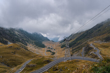 A winding road snakes through the foggy Carpathian mountains, with a steel cable car tower standing prominently in the foreground. The Transfagarasan highway