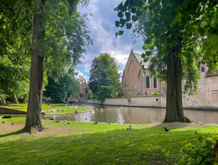 Obraz premium Brugge, Flanders, Belgium - June 25, 2024: Ducks and swans on Minnewater just outside Beguinage building and chapel. Green foliage trees and blue cloudscape