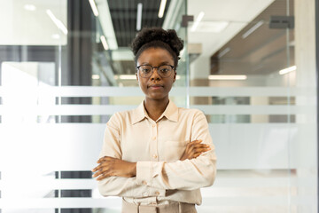 Confident African American businesswoman standing with arms crossed in modern office. Portrays leadership, confidence, and professionalism. Ideal for depicting successful business environment