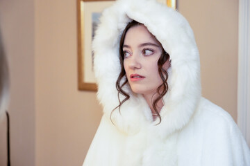 brunette young woman wearing a white fur hooded coat looks to the side with a thoughtful...