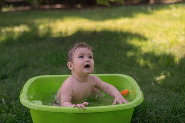 Unhappy boy bathes in a basin outdoors in summer.