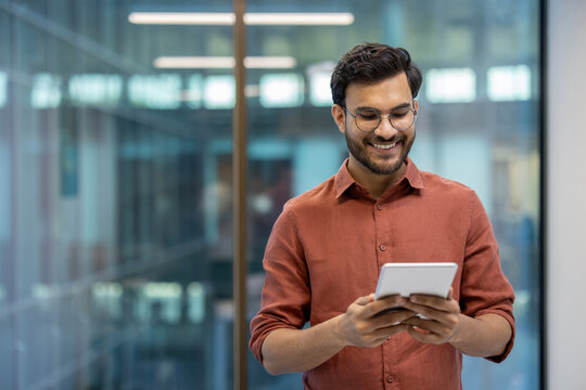 Smiling programmer tests application on tablet in modern office. Casual professional wears glasses and brown shirt, demonstrating focus and tech expertise.