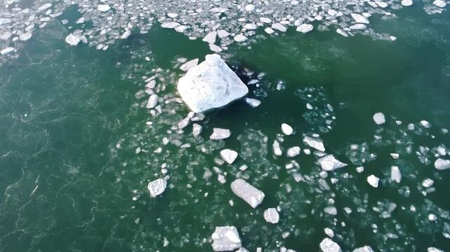 Vue de drone descendante sur un rocher congel&eacute;, envelopp&eacute; d'une pellicule de glace entour&eacute; de morceaux de glaces flottants paisiblement sur l'eau calme verd&acirc;tre. ( Fleuve Saint-Laurent, Qu&eacute;bec. )