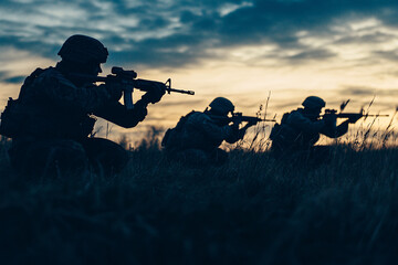Three soldiers crouching in tall grass with rifles aiming at sunset symbolizing military operation and tactical strategy