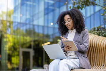Fototapeta premium African American businesswoman participates in an online video course outside. She engages with instructor via laptop, takes notes in notebook uses for video call, enhancing professional skills