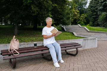 Full length portrait of older woman with short gray hair reading paper book in park, enjoying nature and tranquility, embodying relaxation and wisdom. Elderly female enjoying free time in retirement.