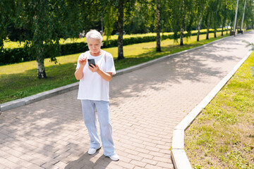 Full length portrait of senior aged woman typing on smartphone while walking in city park. Beautiful gray-haired woman using tapping mobile phone, enjoying online communication.