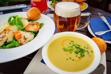 A beautifully arranged dining table featuring a warm bowl of creamy vegetable soup, a fresh salad with grilled chicken, and a chilled beer, creating an inviting atmosphere for a delightful meal.