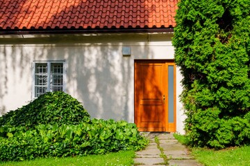 This charming suburban house features a warm brown door, a vibrant red roof, and white walls. The well-kept garden is filled with green plants and colorful flowers, creating an inviting atmosphere for