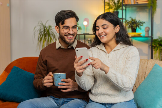 Cheerful young Indian couple experiencing fun, amusement as they browse social media applications together on smartphone. Happy girlfriend boyfriend talking on sofa in home room drinking tea or coffee