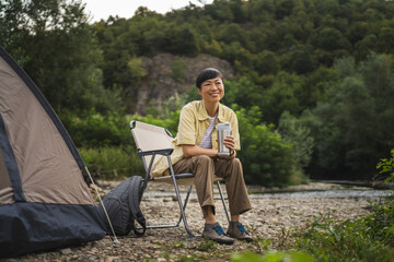 Fototapeta premium Mature japanese woman sit in front of tent and hold thermos