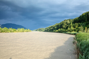 Scenic river flowing through forest with dark storm clouds above, mountains in background, dramatic sky concept