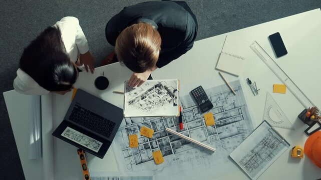Top view of skilled project manager holding project plan and explain house design to architect engineer at meeting table with equipment, ruler, safety helmet and laptop scatter around. Alimentation.