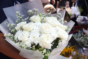 A close-up of an elegant bouquet of white roses, chrysanthemums, and daisies wrapped in white paper, beautifully arranged and fresh, perfect for special occasions