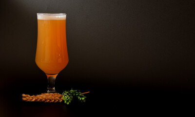 A tall glass of wheat light beer with foam on a black background, next to ears and ripe hop fruits.
