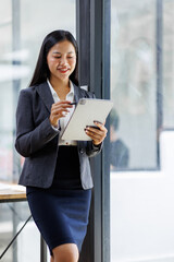 Obraz premium Modern business asian woman in formalwear using digital tablet while standing near wooden desk in an office interior in the office. business people concept.