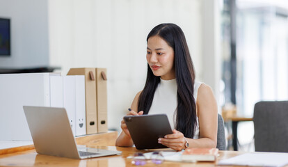 Positive young asian female freelancer in formal suit sitting at table with laptop and browsing tablet while working on project in creative workspace in daylight