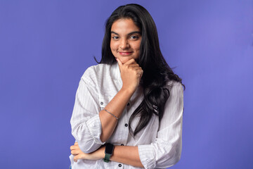 A young woman of Indian descent, wearing a white striped shirt and a black smartwatch, stands with a hand on her chin, exuding confidence and contemplation against a solid purple background