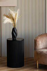 A black table with a vase of flowers on it sits in front of a brown chair. The vase is tall and has...