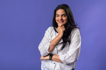 A young woman of Indian descent, wearing a white striped shirt and a black smartwatch, stands with a hand on her chin, exuding confidence and contemplation against a solid purple background