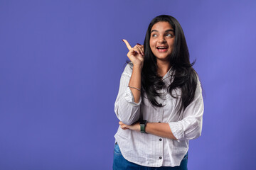 A young woman of Indian descent, wearing a white striped shirt and black smartwatch, stands against a solid purple background, pointing upward as if she just had an exciting idea