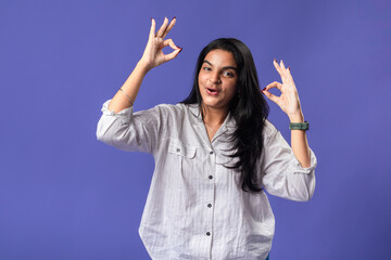 A young woman of Indian descent, wearing a white striped shirt and black smartwatch, stands against a solid purple background, smiling and making an 