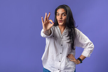 A young woman of Indian descent, wearing a white striped shirt and black smartwatch, stands against...