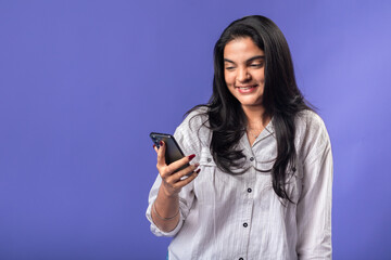 A young woman of Indian descent, wearing a white striped shirt and black smartwatch, stands against a solid purple background, looking at her smartphone,  enjoyment of a message