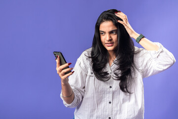 Naklejka premium A young woman of Indian, wearing a white striped shirt and black smartwatch, against a purple background, looking frustrated while holding a smartphone and hand through her hair.