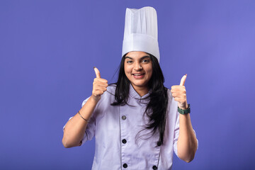 A cheerful Indian woman in a purple chef coat and white chef hat gives two thumbs up, standing against a solid purple background, portraying confidence and positivity in her culinary profession