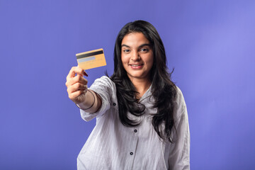 An Indian woman in a casual white shirt holds a credit card toward the camera, smiling confidently. She stands against a purple background, offering a clear view of the card in focus.