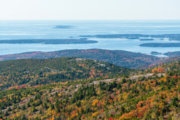 Aerial colorful cliff side downhill mountaintop view of fall foliage and trees in autumn from a hiking area overlooking a bay of blue water facing islands in the distance