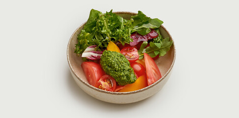 A fresh and vibrant salad is presented in a beige bowl against a clean white background. The salad is made with a mix of fresh greens, ripe tomatoes, and a dollop of vibrant green pesto.