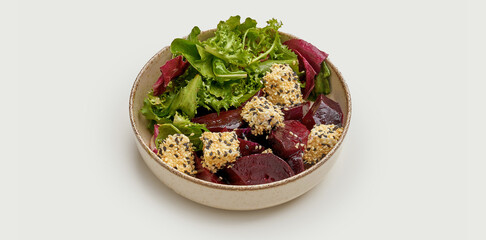 A vibrant and colorful salad is presented in a beige bowl against a clean white background. The salad features roasted beets, fresh greens, and creamy feta cheese topped with sesame seeds