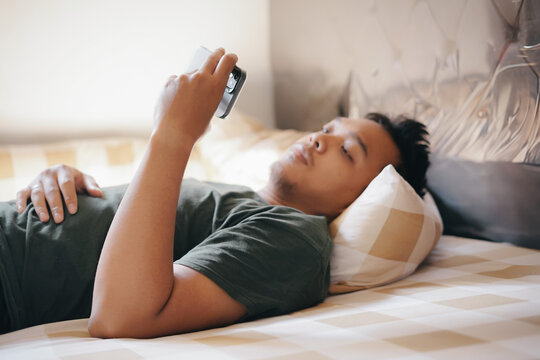 A young man lying on a bed, looking at his smartphone with a thoughtful expression. Relaxed, cozy bedroom setting