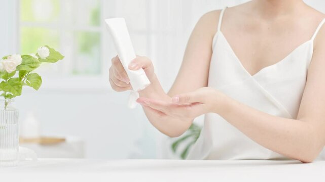 Woman opens an unlabeled white cosmetic tube and squeezes out a small amount of cream onto her fingertips. Bathroom background with bathtub, window and white curtain. Video for product advertising