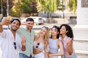 Five friends gather for a selfie in a sunny outdoor setting, smiling and making peace signs, capturing a fun moment together with greenery in the background. 
