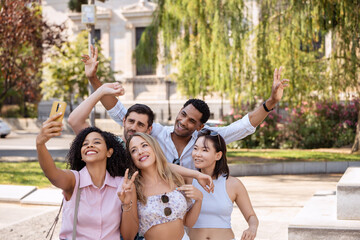 A cheerful group of five friends taking a selfie in a sunny park, smiling brightly and posing with peace signs, enjoying a fun, carefree moment together. 