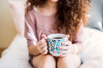 Morning cup of coffee. Beautiful young woman with curly hair sitting on bed in her room, drinking hot coffee.