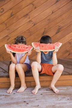 Boy and girl hiding faces with watermelon slices and sitting at porch