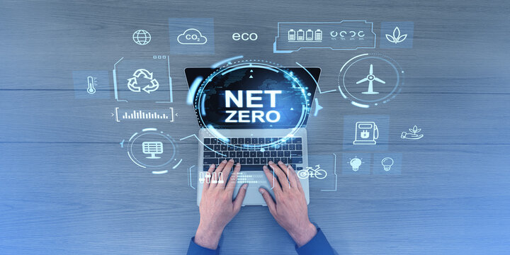 Hands typing on a laptop displaying NET ZERO surrounded by eco-friendly icons like recycling, wind turbine, and plant on a wooden background. Concept of sustainable energy and ecology