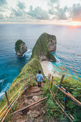 A young man stands on a rock by the beach against a colorful sunset sky with a beautiful view of the turquoise sea and clouds at sunset at Kelingking Beach, Nusa Penida, Bali. © Balnyes Visuals