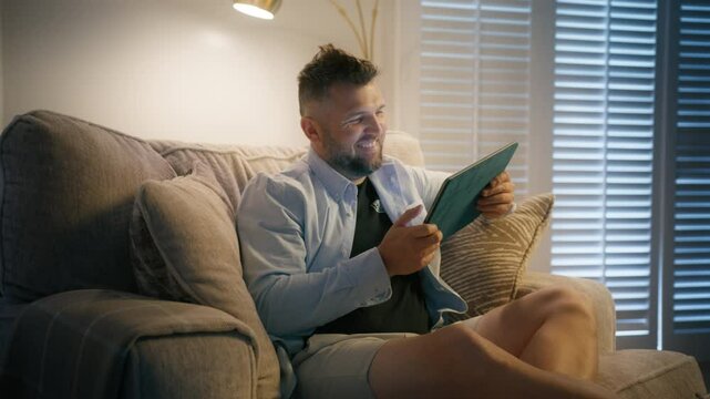 A cheerful man sits comfortably on a cozy couch, fully focused on his tablet, enjoying a relaxed evening surrounded by warm lighting that creates a nice ambiance of contentment