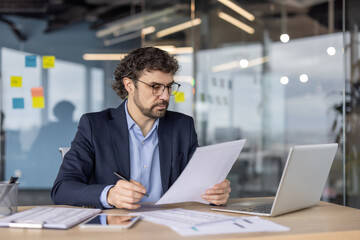 Businessman in formal suit focused on paperwork at office desk. Concentrated on financial documents, using pen and laptop for detailed analysis. Modern workspace reflects professional environment.