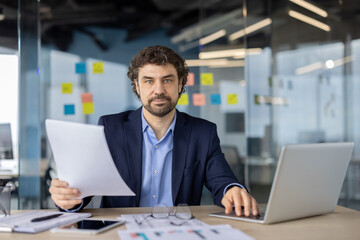 Fototapeta premium Serious businessman working in modern office holding document. Thoughtful professional analyzing information while using laptop. Focused on paperwork, enhancing productivity